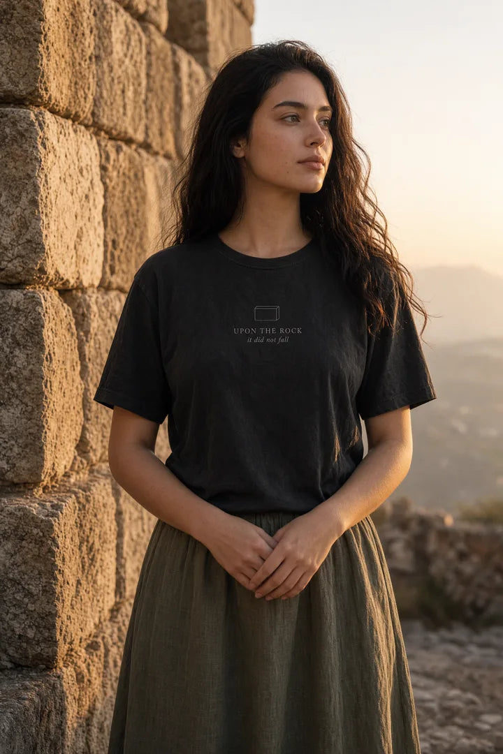 Woman with clasped hands by a chapel wearing a washed black Upon the Rock cornerstone tee.